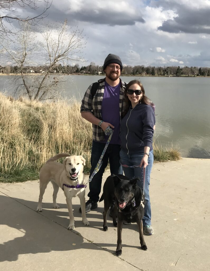 Two people and two dogs standing in front of a lake