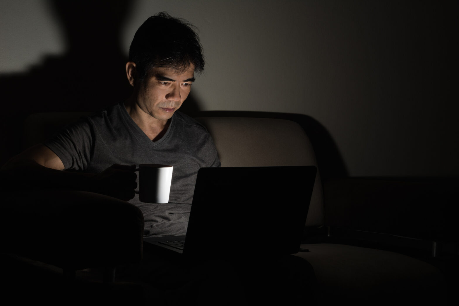 A man working on his laptop at night, sitting on a couch and holding a white mug